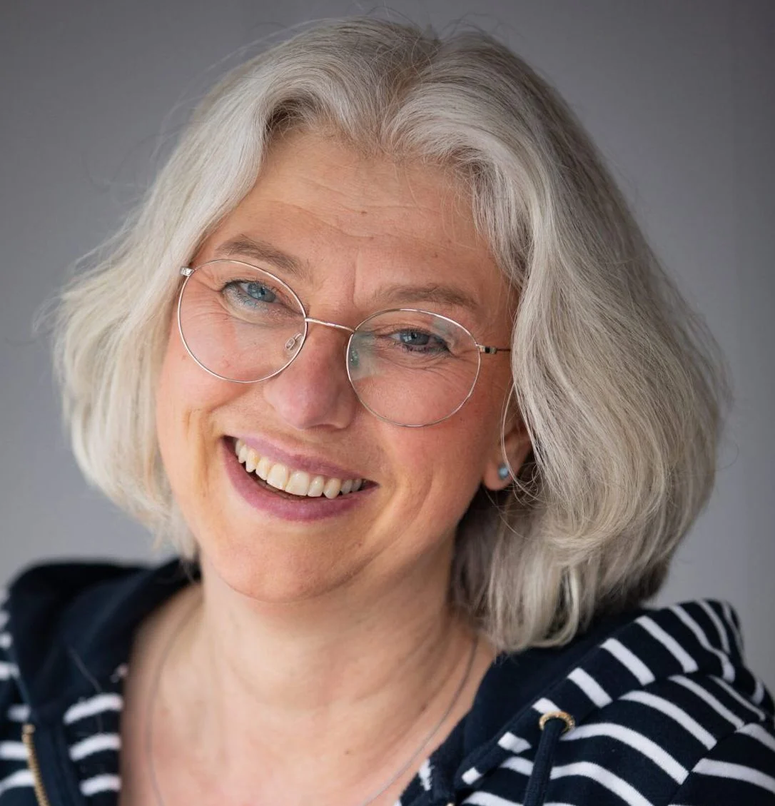 Smiling woman with shoulder-length gray hair wearing glasses and a striped hoodie, set against a neutral background