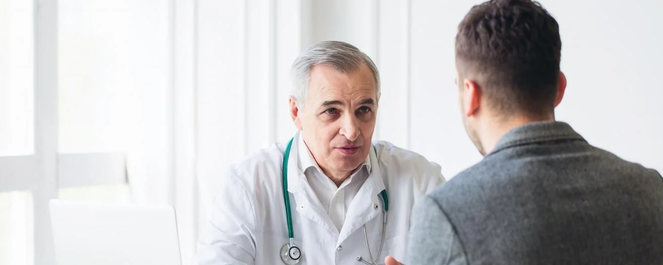 A doctor in a white coat speaks intently with a male patient in a bright, modern office setting