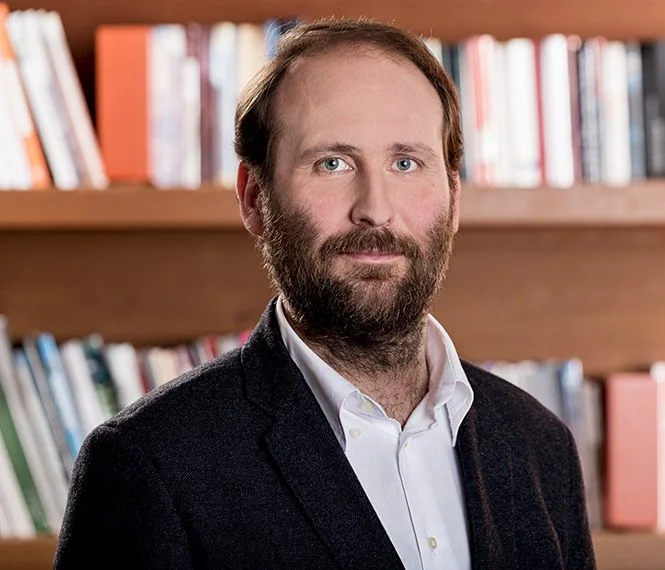 A man with a beard poses confidently in front of a wooden shelf filled with books, wearing a dark blazer and white shirt