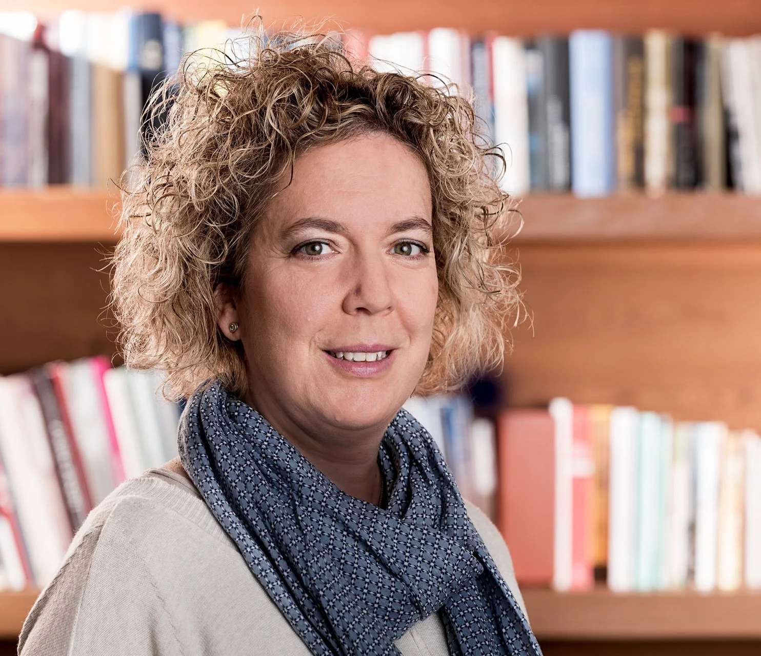 A woman with curly hair wears a blue patterned scarf, smiling against a bookshelf filled with colorful books