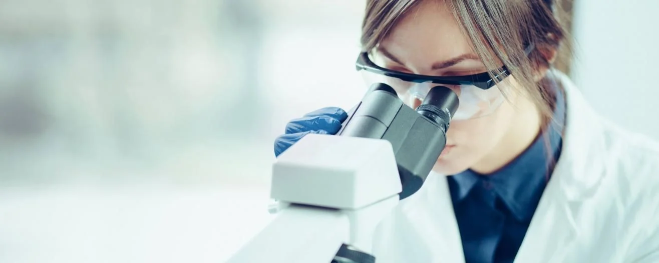 A focused woman wearing a lab coat and goggles examines a specimen through a microscope in a bright laboratory