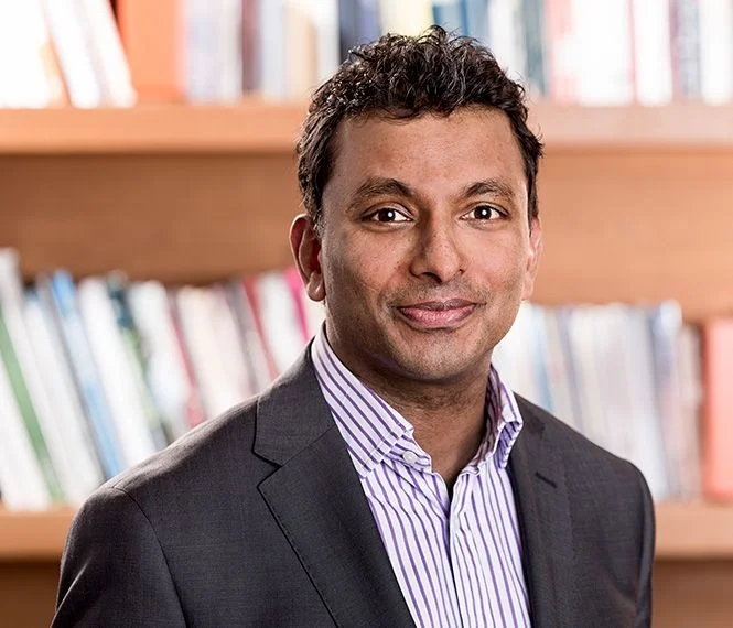 Smiling man in a gray suit with a striped shirt stands in front of a bookshelf filled with books