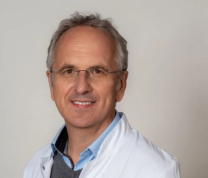 Man with gray hair, glasses, and a white coat smiles warmly against a plain light background