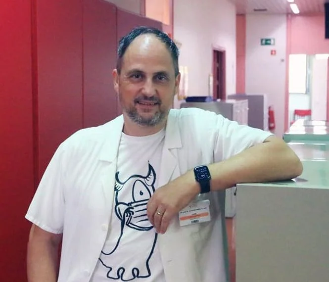 A smiling man in a white lab coat poses casually with one arm resting on a counter in a brightly lit hallway