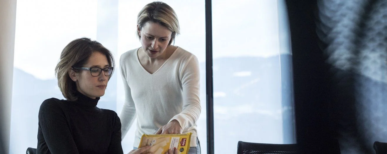 A woman in a black turtleneck listens as another woman in white hands her a document, with mountains visible through a window