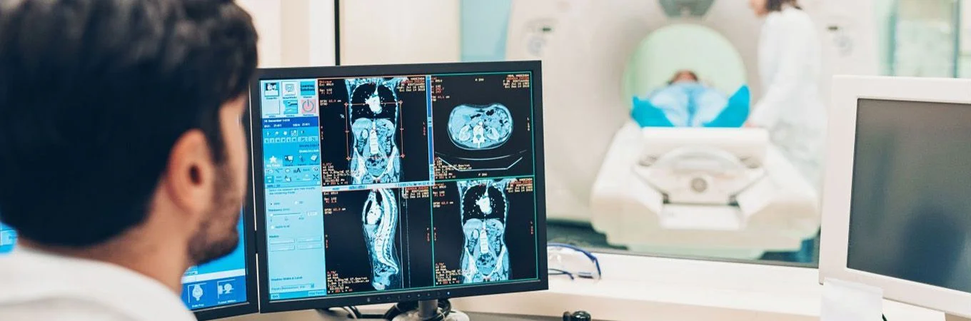 A medical professional analyzes CT scan images on a monitor while a patient lies on the scanning machine behind