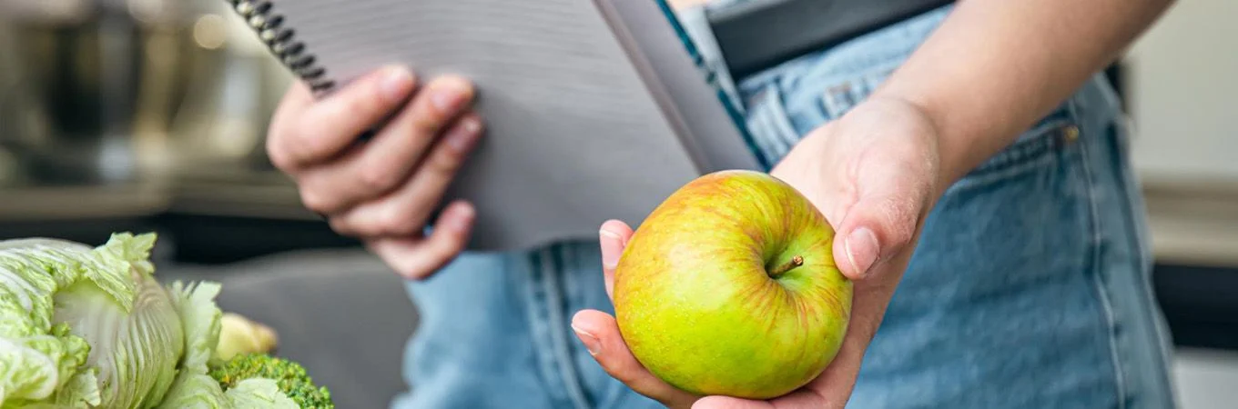 A person holds a green apple in one hand while a notepad is clasped in the other, surrounded by fresh vegetables
