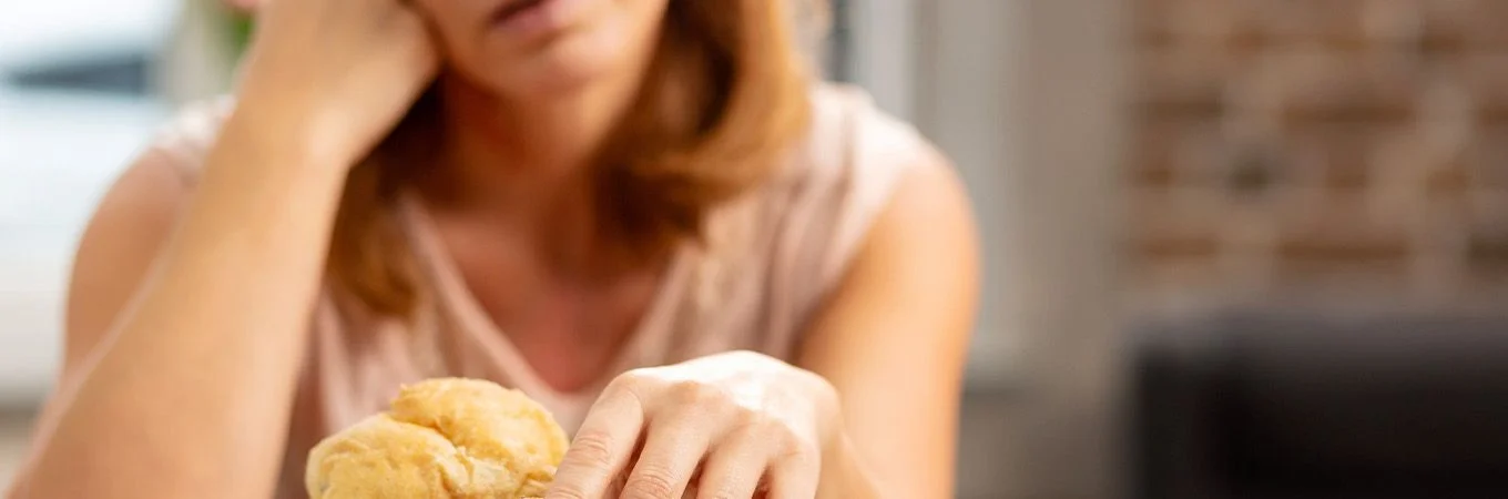 A woman rests her head on her hand, staring pensively at a bun on the table, reflecting feelings of discontent or frustration