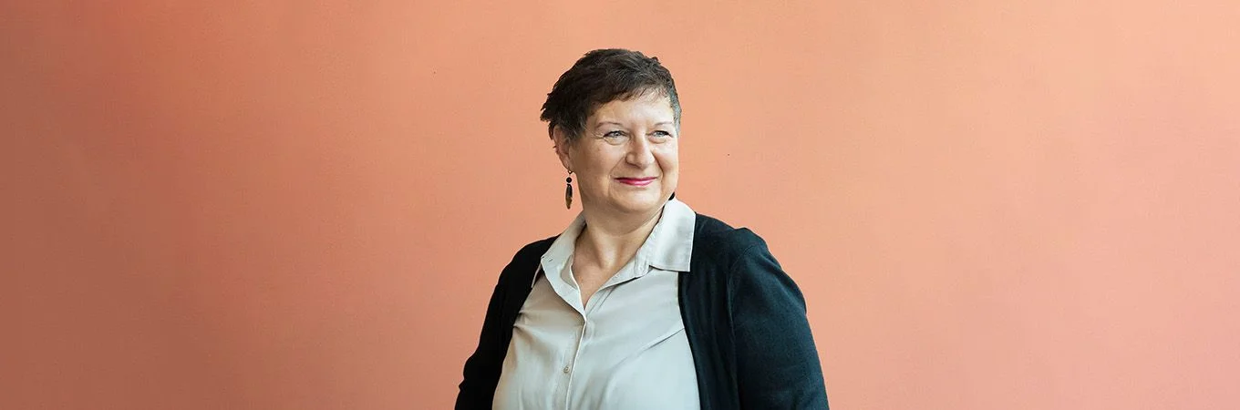 A woman with short hair and earrings smiles, wearing a light blouse and black cardigan against a soft orange backdrop