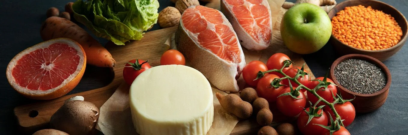 A wooden cutting board displays a variety of fresh foods: salmon, tomatoes, lettuce, grapefruit, lentils, mushrooms, and cheese