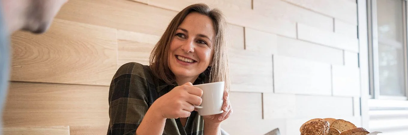 Smiling woman in a plaid shirt joyfully holds a cup of coffee, with freshly baked bread on the table beside her