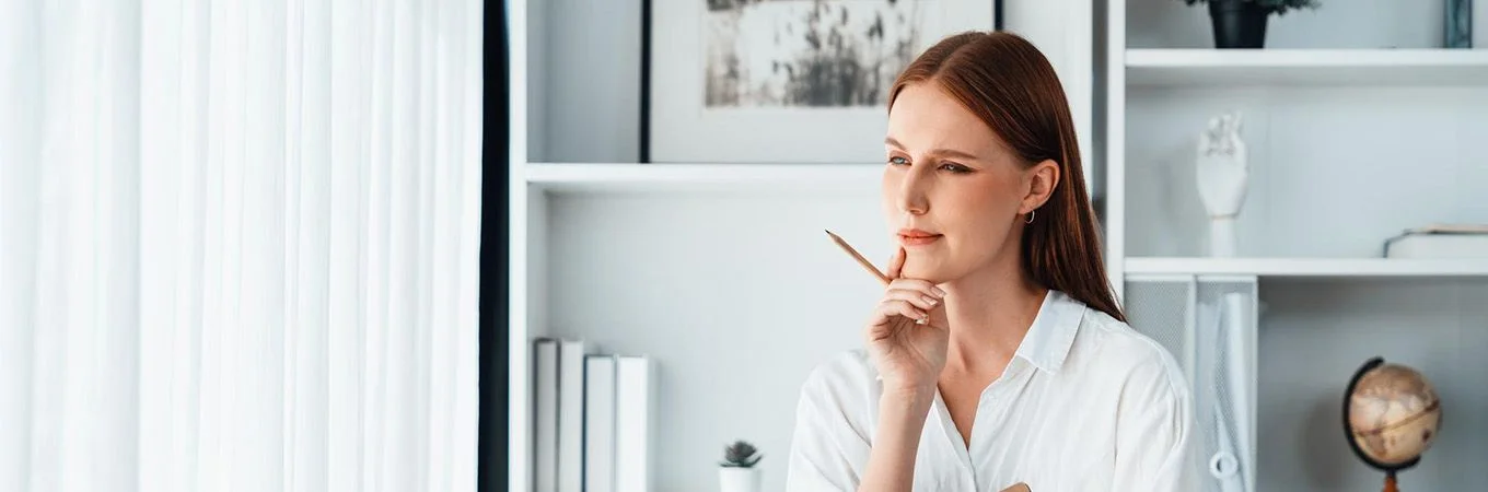 A woman with long red hair thoughtfully holds a pencil, surrounded by shelves and soft white curtains in a modern workspace
