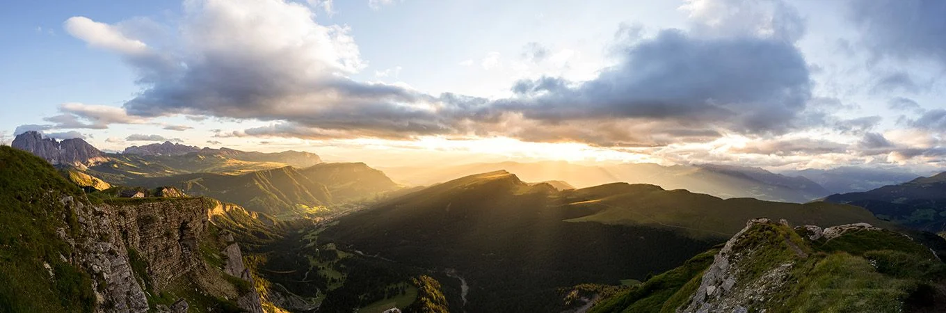 Sunset illuminates a vast mountainous landscape, with rays piercing through clouds over lush valleys and rugged cliffs