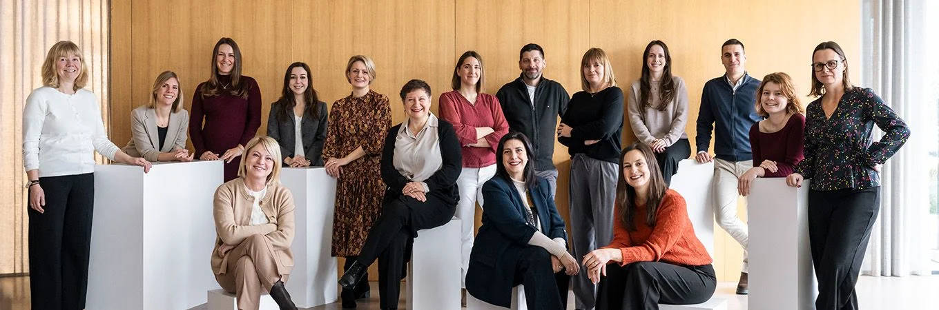 A diverse group of 20 adults poses in a modern indoor setting, smiling and standing around white pedestals against a wood backdrop