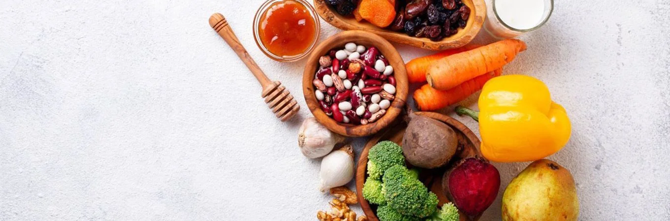 A variety of fresh vegetables, beans, honey, and dried fruits arranged on a textured surface, showcasing vibrant colors and textures