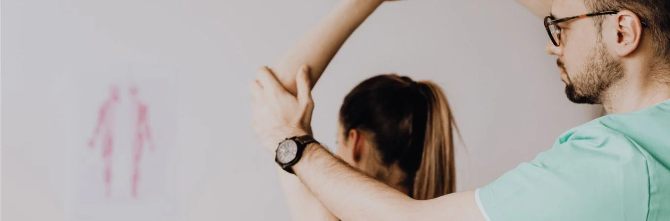 A healthcare professional assists a patient by stretching her arm in a clinical setting with anatomical charts on the wall