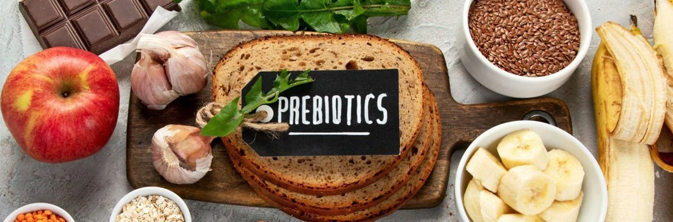 Sliced bread labeled "Prebiotics," surrounded by fruits, vegetables, garlic, chocolate, and various grains on a wooden board