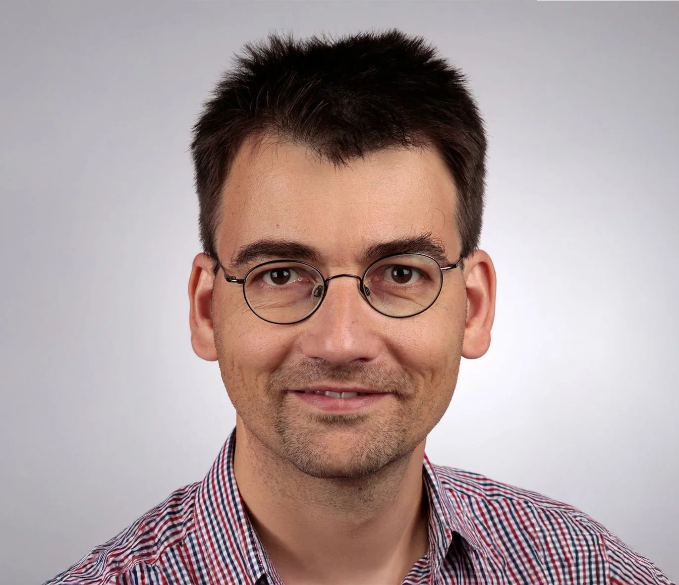 Man with short, dark hair and glasses smiles, wearing a checkered shirt against a light gray background