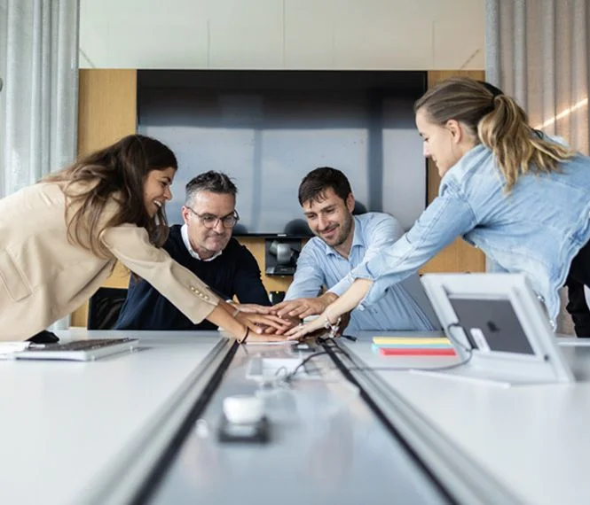 Four diverse professionals join hands in a collaborative gesture at a modern conference table, with screens in the background