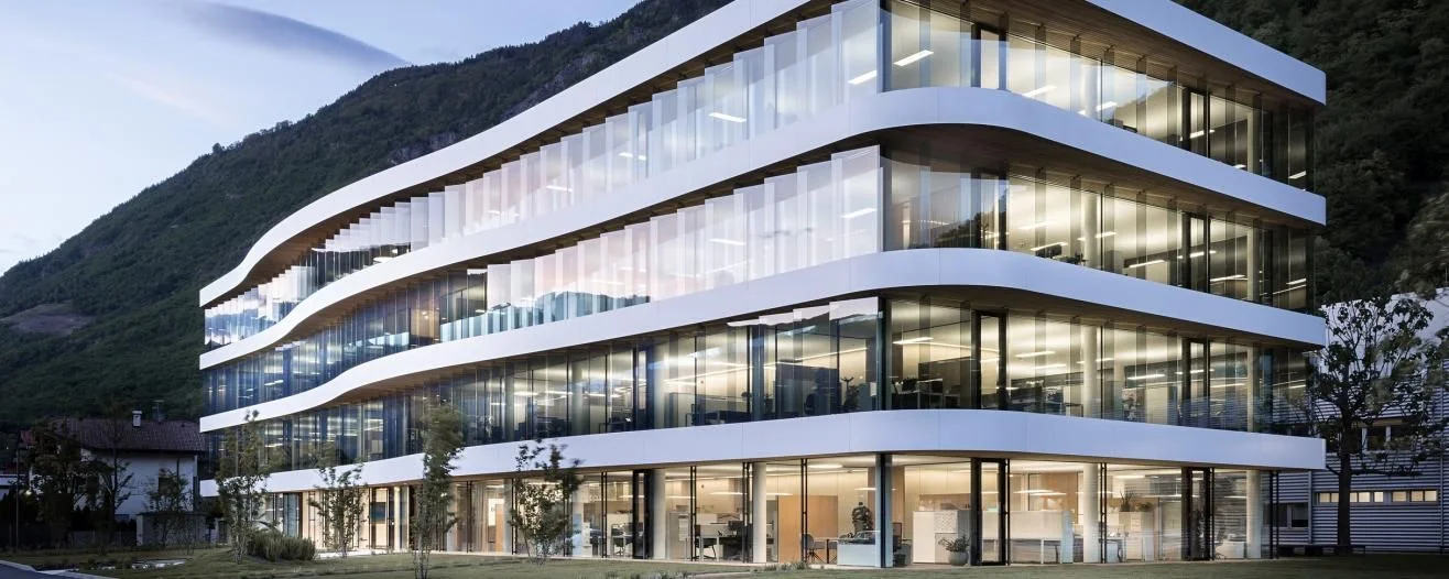 Modern, wavy glass building set against a mountainous backdrop, featuring landscaped grounds and bright interior workspaces