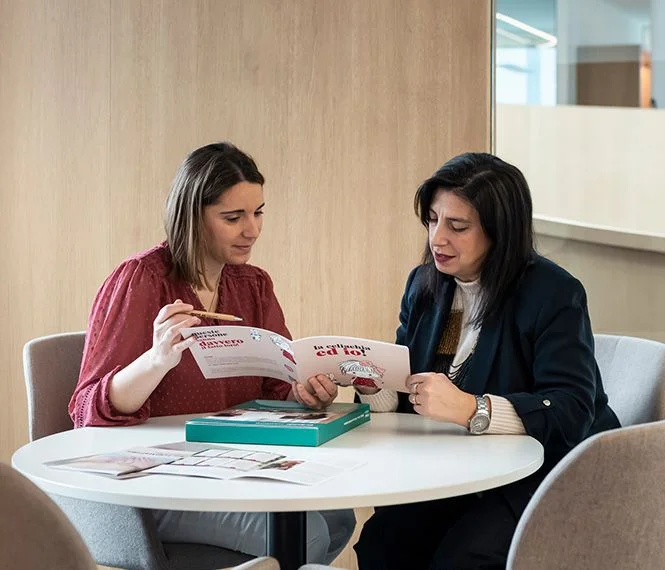 Two women discuss and review materials at a round table, with a light wooden backdrop and a green box between them