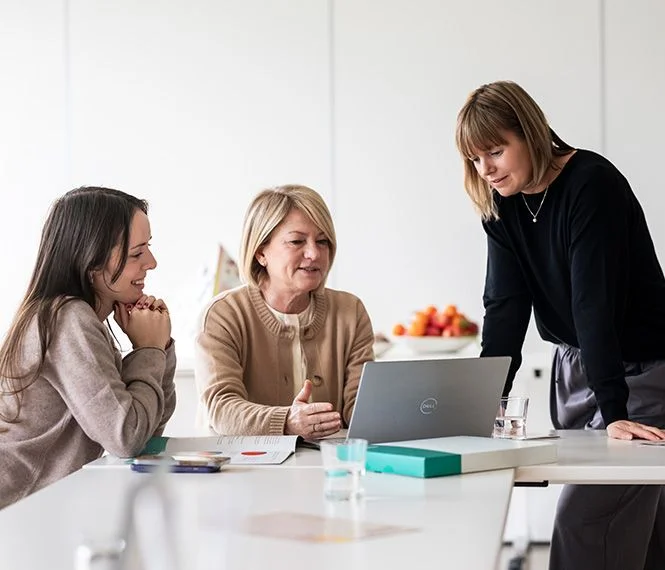 Three women gather around a laptop in a bright meeting room, discussing ideas, with fruits visible in a bowl nearby