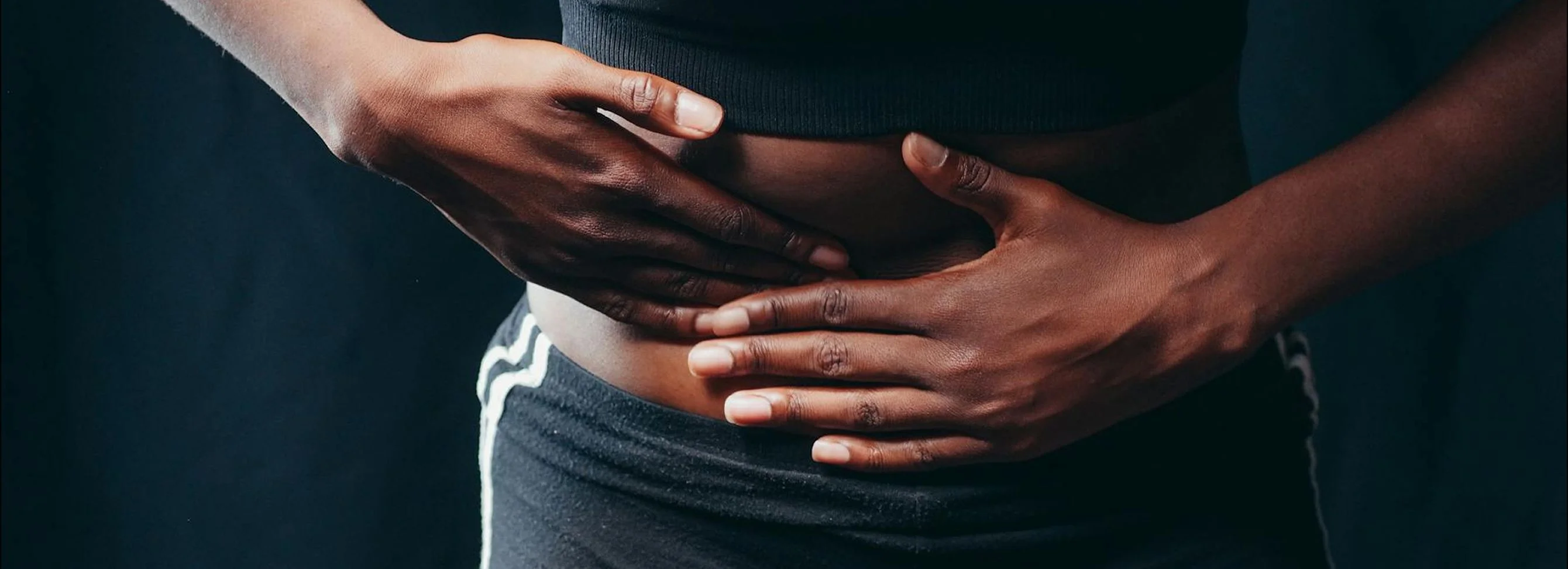 Hands resting on a bare midsection, conveying discomfort, against a dark background. Soft lighting highlights the skin tones