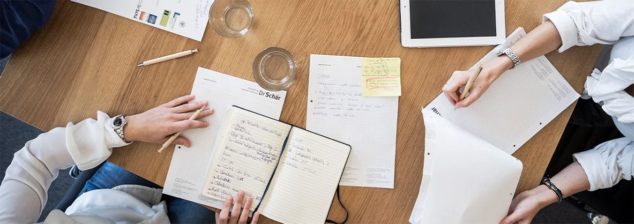 Multiple hands engaged in writing and note-taking around a wooden table, featuring papers, notebooks, and an iPad