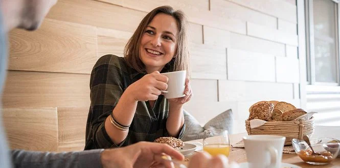 A smiling woman holds a cup of coffee, enjoying breakfast in a cozy setting with fresh bread and juice on the table